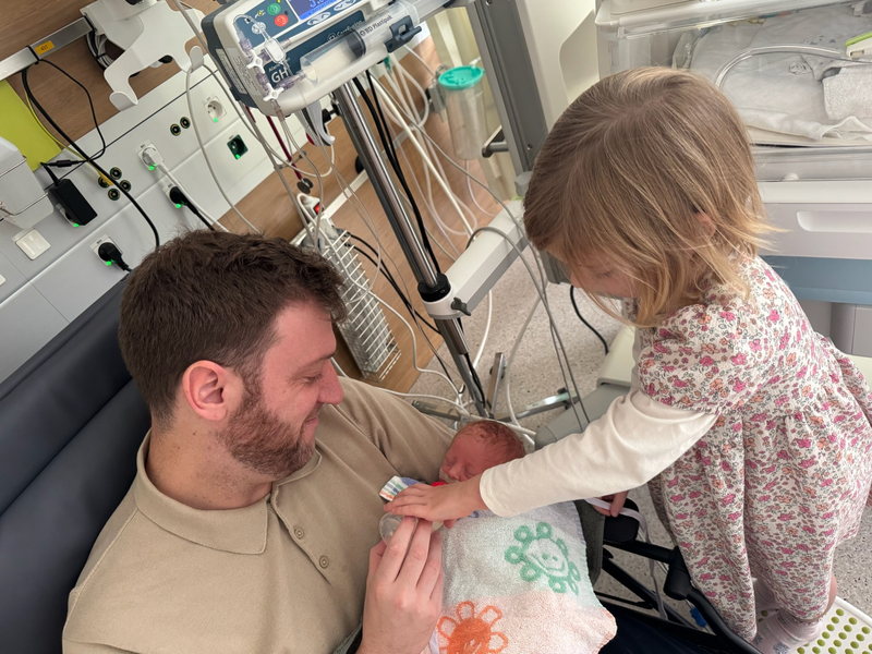 Loïc, pasgeboren en ingebakerd, in de armen van zijn papa terwijl zijn grote zus zijn handje vasthoudt.